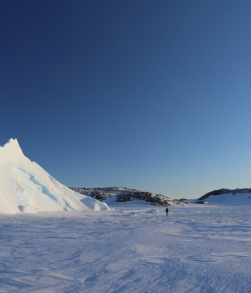 Expanse of sea-ice with pyramid ice berg sticking up out of it, two people walking across sea-ice towards the burg. Bright blue sky above, rocky hills in background
