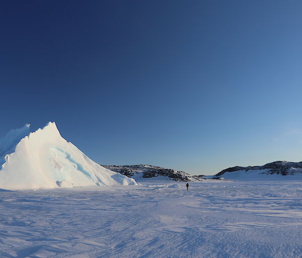 Expanse of sea-ice with pyramid ice berg sticking up out of it, two people walking across sea-ice towards the burg. Bright blue sky above, rocky hills in background