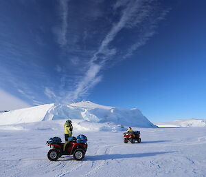 Foreground, man standing beside quad bike on the sea-ice. Distance another quad bike with stranded burg, and blue sky above