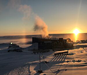 Sunrises over the plataue in the distance, in the forground the blue main powerhouse building emitting steam from the chimney and the snow covered ground has an orange sunlit tint