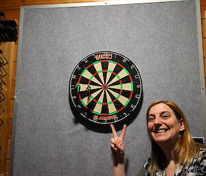 Woman in foreground grining and giving Victory symbol with hand in front of darts board with three darts in the 11 section