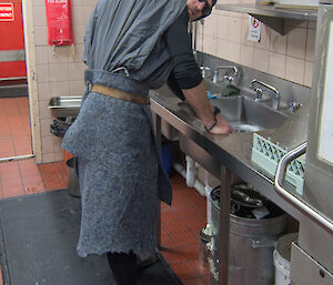 Man dressed as hunchback in grey felt is standing at sink washing dishes