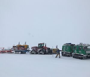 Man stands at Antarctic circle sign with vehicles lined up behind — green Hägglunds on right and large tractor with two trailers attached on left, on middle trailer are two yellow snow blowers.