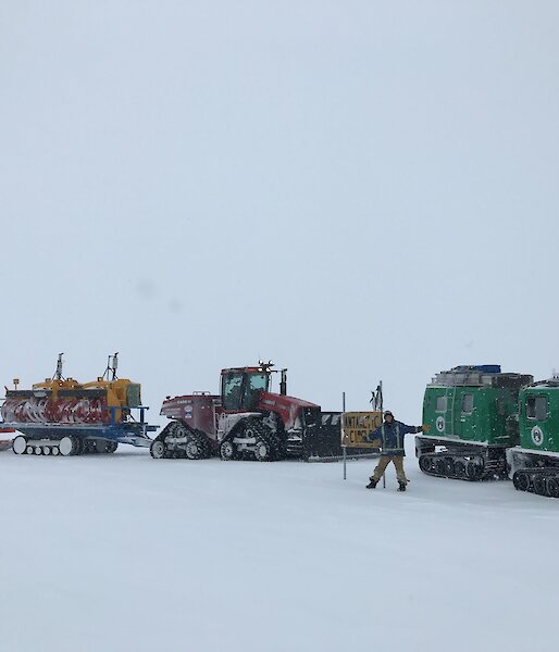 Man stands at Antarctic circle sign with vehicles lined up behind — green Hägglunds on right and large tractor with two trailers attached on left, on middle trailer are two yellow snow blowers.