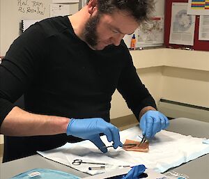 Man sitting at table suturing a square piece of pig skin, wearing blue surgical gloves.