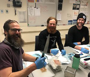 Three men sit at table with whiteboards behind. They wear blue surgical gloves and in front of them are clothes laid with suturing equipment. Man closest on left is holding up a chef’s knife and smiling.