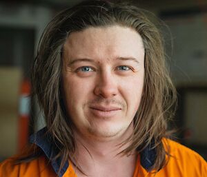 Headshot of man in hi-vis shirt with long shoulder length hair and a partially grown moustache and goatee.