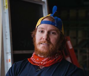Headshot of man in blue shirt with red bandana around neck, wearing ball cap backwards that has helicopter propellors on top, with a short red beard.