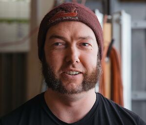Headshot of man in black t-shirt with maroon beanie, with mutton chops beard.