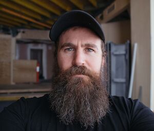 Headshot of man in black t-shirt wearing black ball cap with full dark brown beard with tinges of grey.