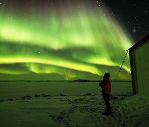 Man standing beside building on right, with bright green aurora spread across the sky above.