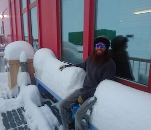 Man sits in centre of bench seat with mounds of snow to mid-chest height either side.