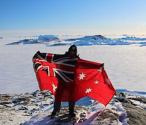 Man standing on rocky snow-covered outcrop with sea ice and snow covered islands in background, holding up an Australian Merchant Navy ensign.