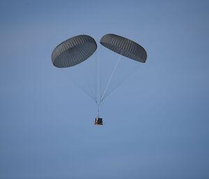box of cargo suspended below two open khaki parachutes, blue sky background