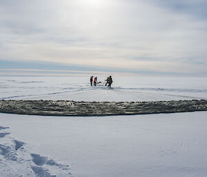 Khaki parachute spread across the snow covered ground, lines extend out into the distance where a grup of people stand