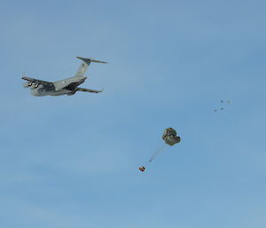 C17 flies overhead, back ramp is down, large box attached to just opening parachute is dropping from the plane, blue sky background