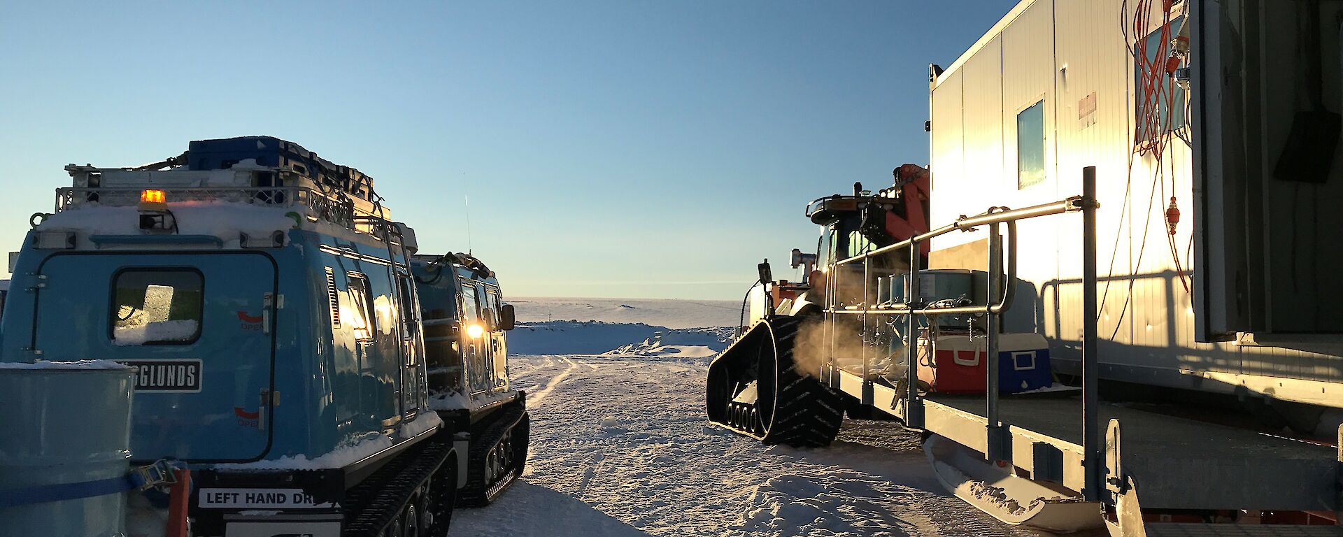 Looking from behind at blue hagglunds with tailer and tractor with accommodation van, both pointing towards plateau in distance with clear blue sky above