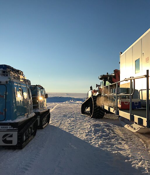Looking from behind at blue hagglunds with tailer and tractor with accommodation van, both pointing towards plateau in distance with clear blue sky above