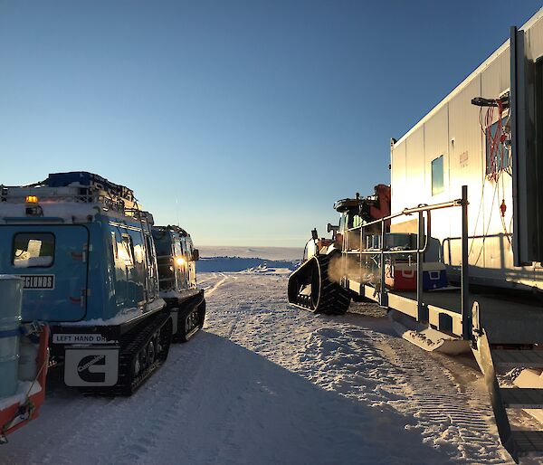 Looking from behind at blue hagglunds with tailer and tractor with accommodation van, both pointing towards plateau in distance with clear blue sky above