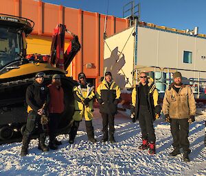 Group of six men standing facing camera, wearing cold weather gear, behind is tractor and accommodation van then orange building