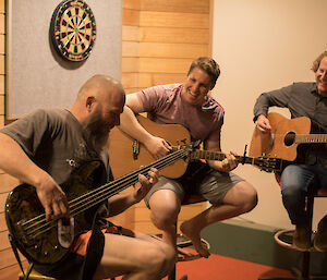 Three men sitting on bar stools, two with guitars and one with bass. Behind wooden panelled wall with darts board.