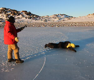 Man lies face down on ice lake, while second man looks on taking photo