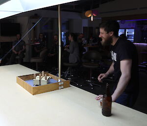 Game of skittles sits on large wooden table, man in black t-shirt plays the game with a bottle of beer on table beside