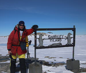 Man stands in red parker, leaning on “Shirley Island” metal sign. Background is snow covered valley with blue sky above.