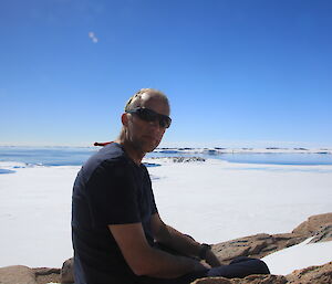 Man in blue t-shirt and sunglasses sits on rocky outcrop with ice covered bay behind and blue sky above