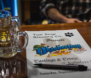 Poster on wooden bar reading “Two Dogs Brewing Co presents Oktoberfest Tastings and Trivia at Splinters 2”, a pen rests over the poster and two beer glasses are beside it