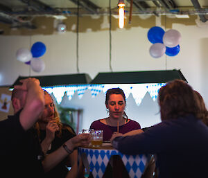 Five people sitting around a bar table answering trivia questions and drinking beer. Blue and white bunting around table and blue and white balloons hanging from ceiling