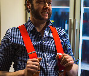 Man sitting on bar stool wearing brown shorts, blue and white checked shirt and red braces