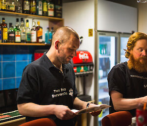 Man standing behind bar with shelves of bottles and bar fridge behind stands and reads from A4 paper, wearing 2 Dogs Brewing Co t-shirt