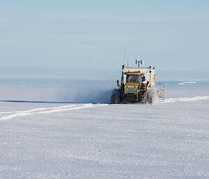 Snow covered slope extending to sea in distance. Tractor pulling shipping container on sled is travelling along tracks in the snow