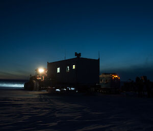 View of traverse camp at dusk, with lights on above the tractor and in the van, behind the hagglunds is parked beside the van
