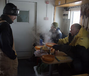 Men sitting and standing around table inside van, still wearing cold weather gear, one sitting on right is serving out spaghetti which is steaming in the cold