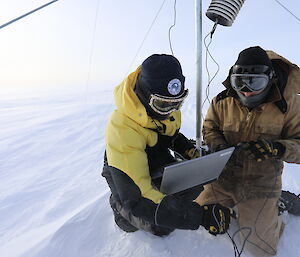 Two men sitting at base of AWS tower looking at laptop, dressed in cold weather gear including beanies and goggles