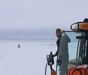 Driver of tractor stands at door and smiiles back to camera, in the distance hagglunds in driving down the slope towards the sea and Casey station