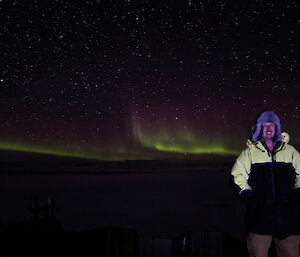 Man to left of picture in cold weather clothing including fur hat, at night with green Aurora in background