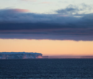 Looking across open water to tounge of glacier at sunset, glacier highlighted in pink light, with orange and pink on horizen with grey clouds above