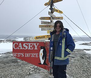 Man in work clothes and blue high vis jackets stands beside the Casey sign which displays distances to multiple world wide locations
