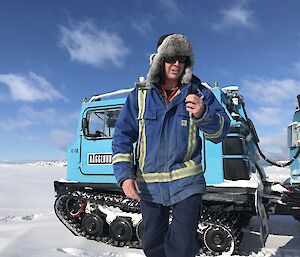 Man in centre of picture, in blue high vis jacket and wearing fur hat and sunglasses. In background front compartment of blue hagglunds vehice. Snow covered ground and blue skies above.