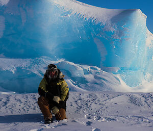 Man kneels in snow in front of bright blue stranded iceberg