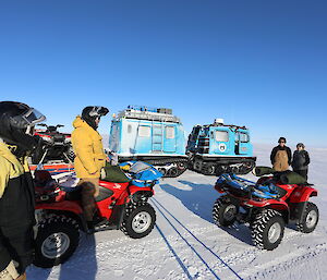 Blue hagglunds with two quads on trailer behind, two quads on ice in front, with four people standing beside, blue sky above