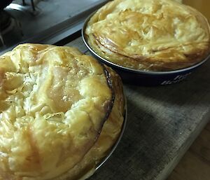 Picture of two pies, pastry on top sitting in large flat tin, sitting on wooden bench