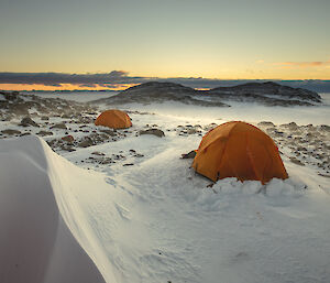 Two polar dome orange tents erected amongst snow and rocks, in the distance a ice covered channel then two rocky hills, at sunset