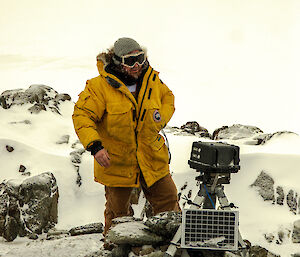 Man in winter gear, including yellow goose down coat, stands beside camera box on metal stand with solar panel in front. On rocky snow covered ground with sea ice behind in distance