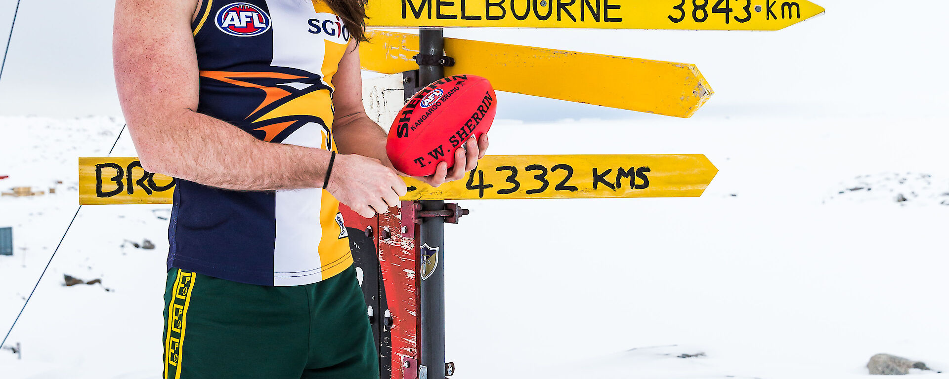 Man stands in AFL top and short in big boots and beanie and holding football beside Casey sign that indicates Melbourne is 3843km away