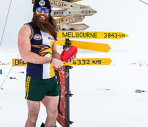 Man stands in AFL top and short in big boots and beanie and holding football beside Casey sign that indicates Melbourne is 3843km away