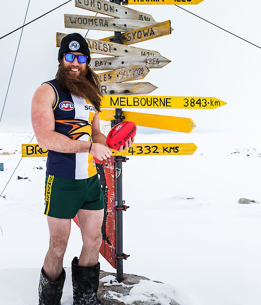 Man stands in AFL top and short in big boots and beanie and holding football beside Casey sign that indicates Melbourne is 3843km away
